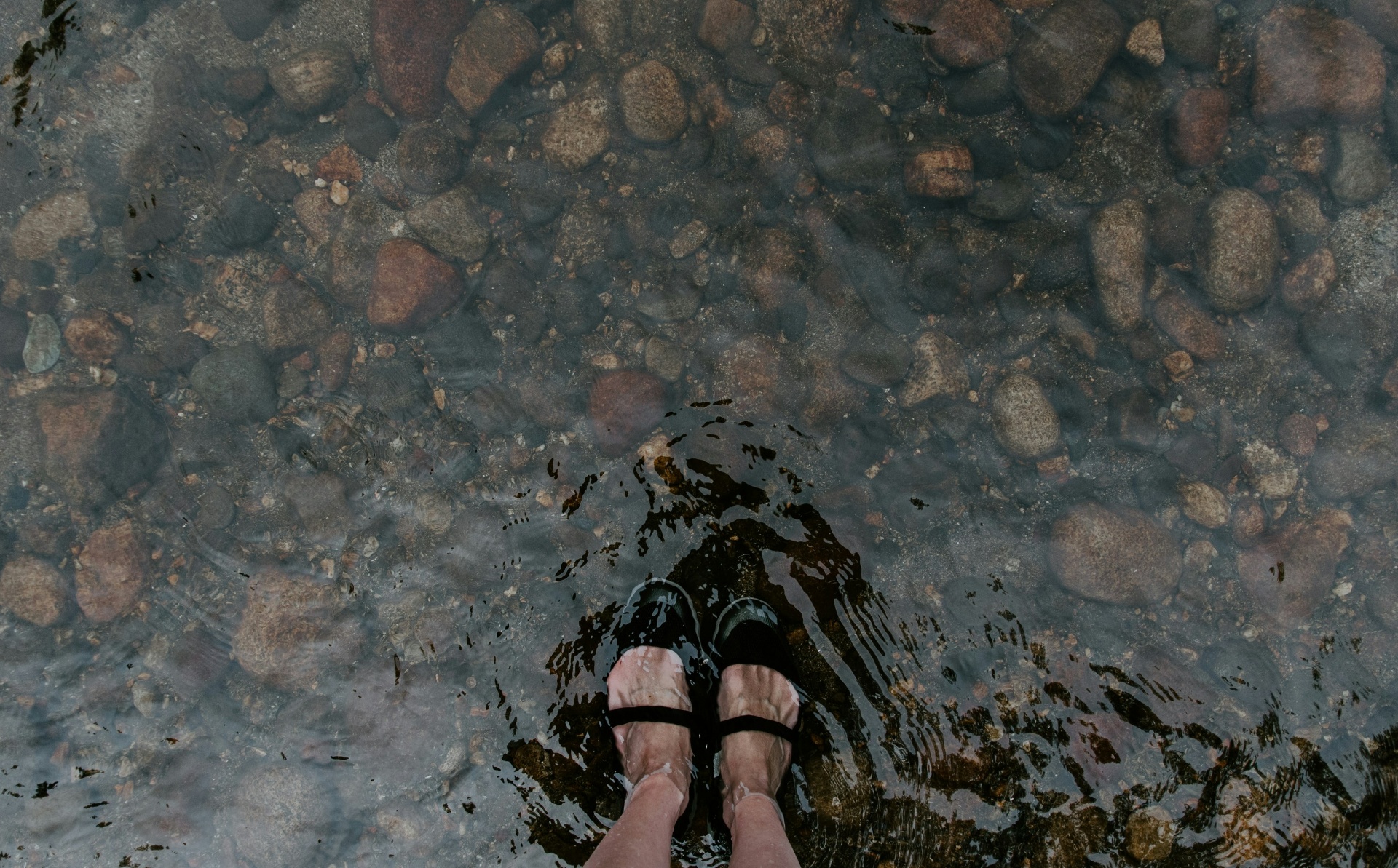 Top-down view of feet in black sandals standing in a clear, shallow rocky stream